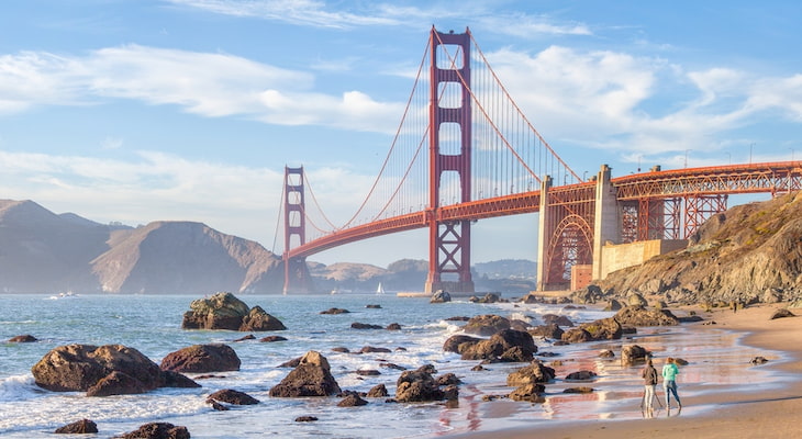 view of the Golden Gate Bridge from Baker Beach in San Francisco. 3 beachgoers walk the sand in the foreground, with the bridge looming in the background